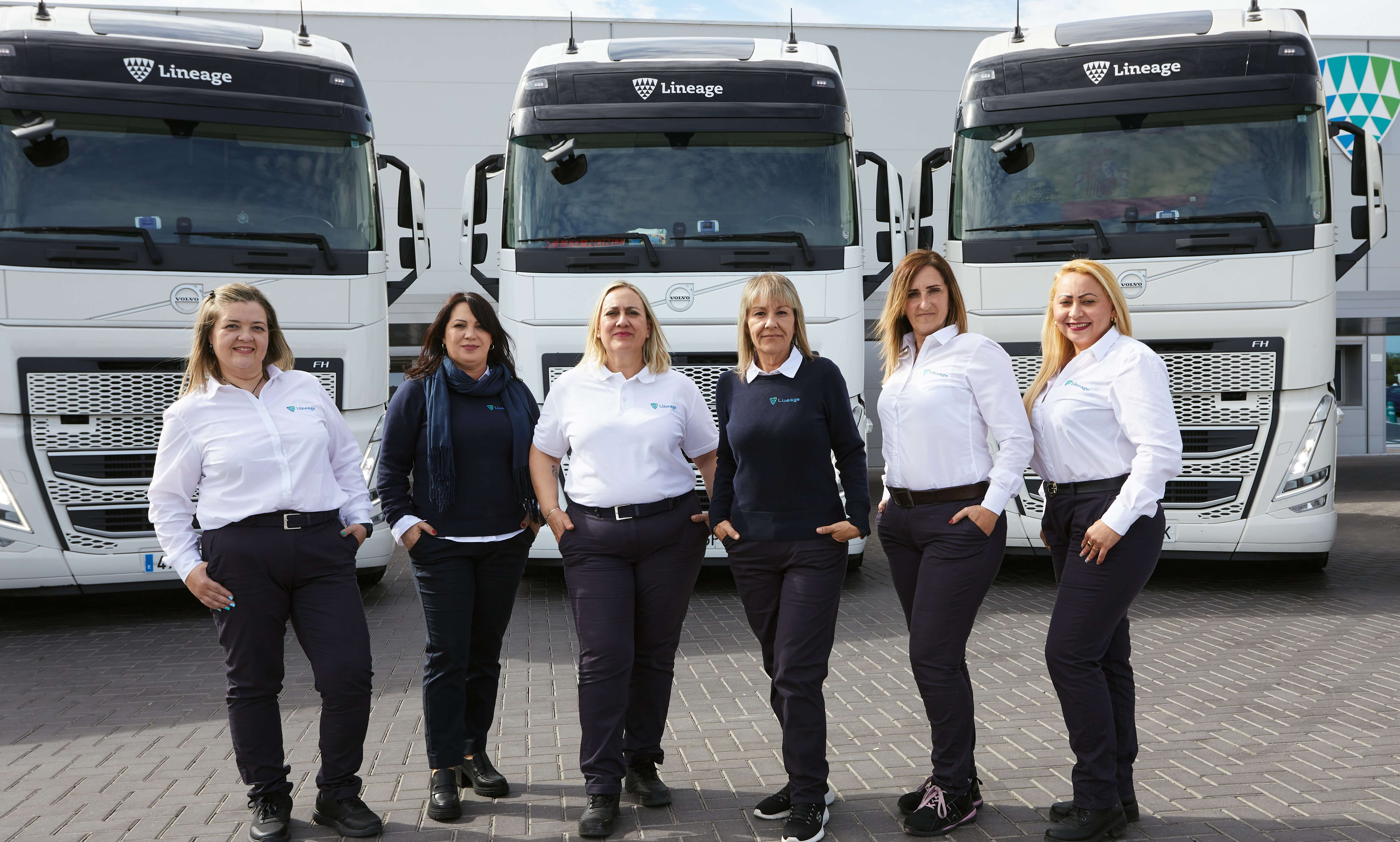 Five women drivers in white shirts and dark trousers standing confidently in front of Lineage logistics HGVs.