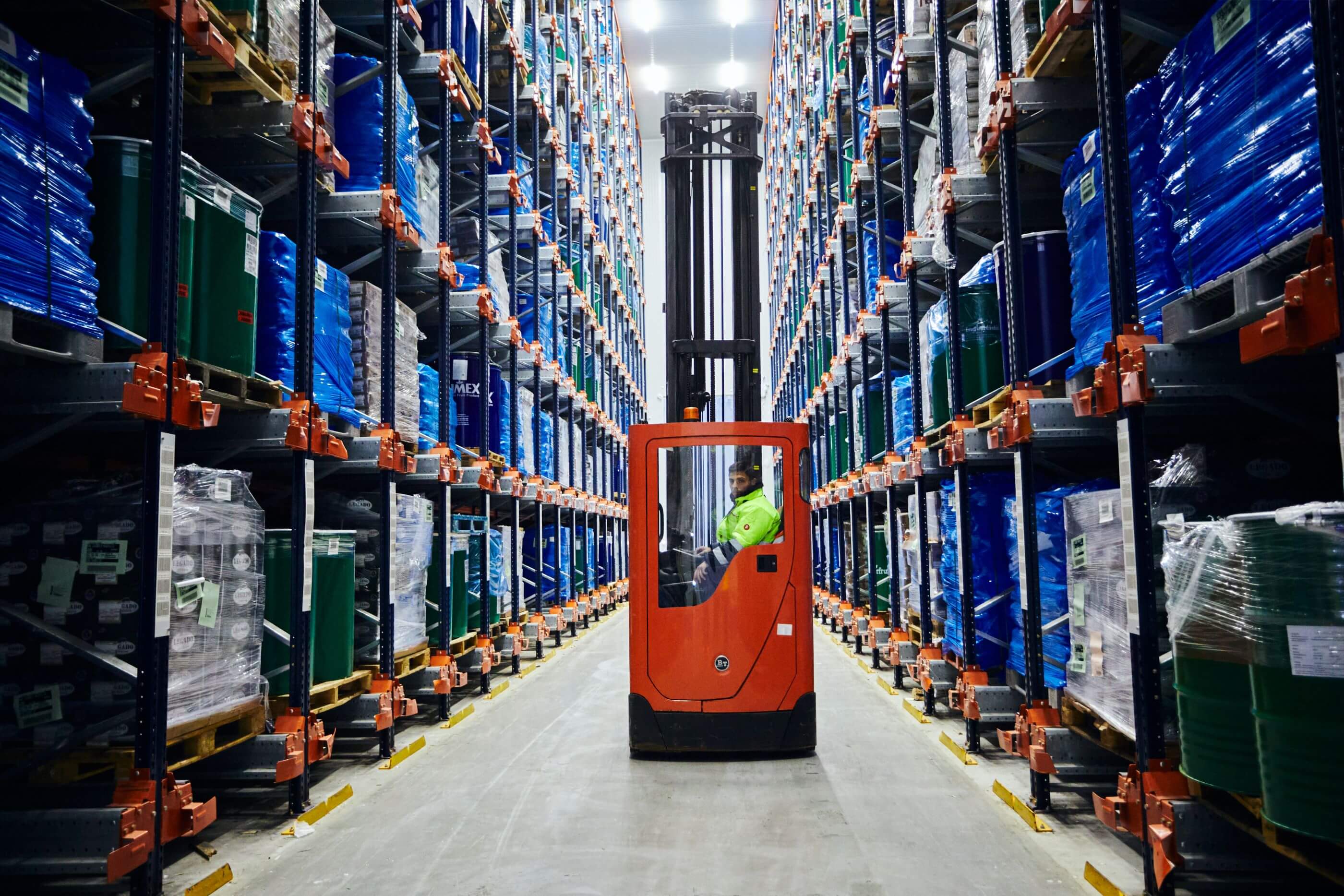 A forklift operator navigating through tall storage racks filled with goods in a warehouse.