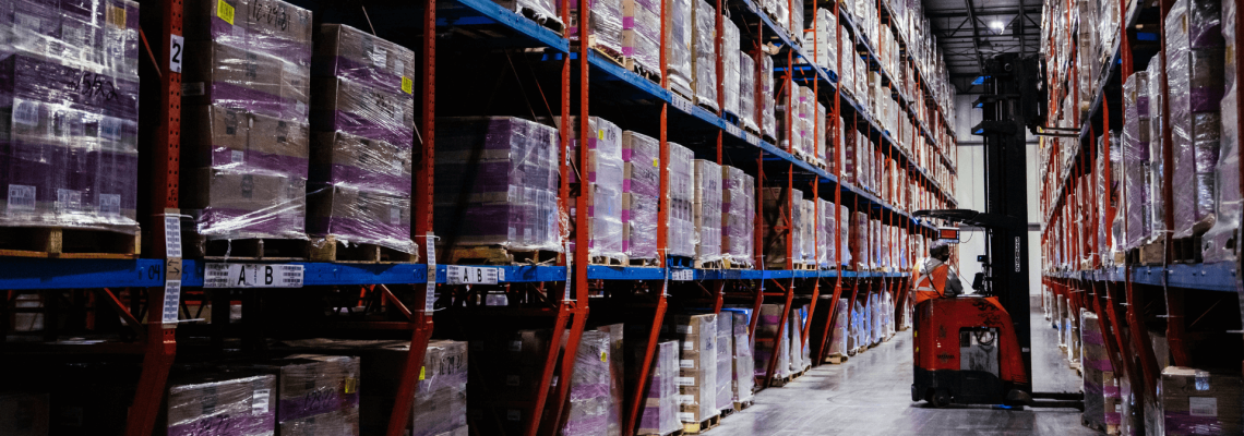 Interior view of a cold storage warehouse with high shelves packed with pallets wrapped in purple plastic, under dim lighting. A forklift is operated by a worker in the central aisle.